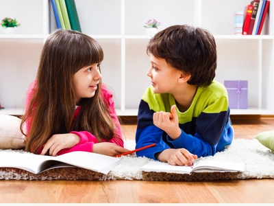 Two young students lie on the floor with open books, facing each other and talking, showing engaged conversation and peer connection during a learning activity.