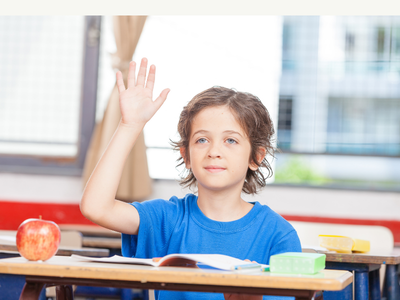 Elementary student sitting at a desk with a hand raised, showing readiness to participate and growing confidence during a classroom lesson.